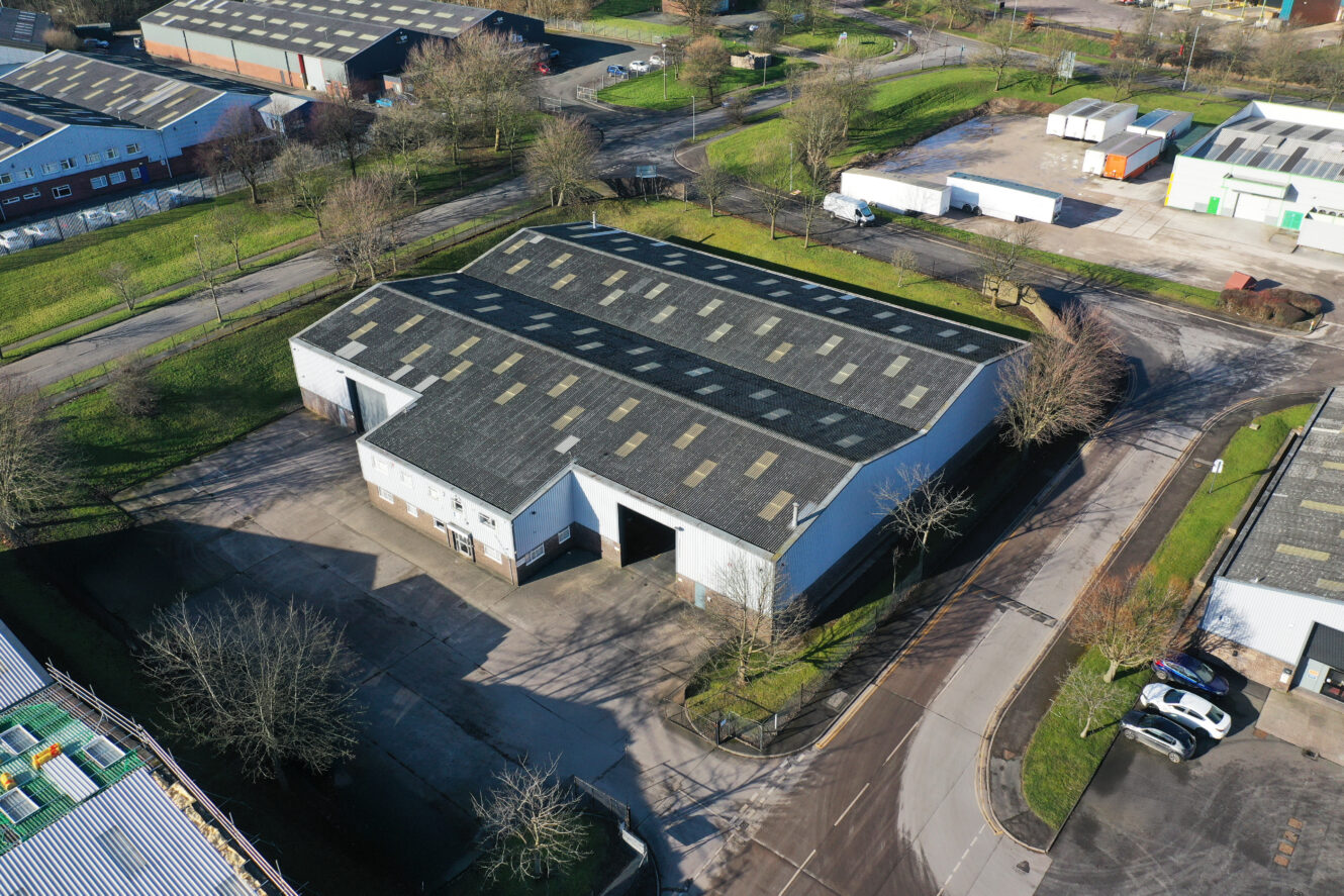A large rectangular industrial warehouse with skylights on the roof, surrounded by paved roads, grass, and parked vehicles in a business park.