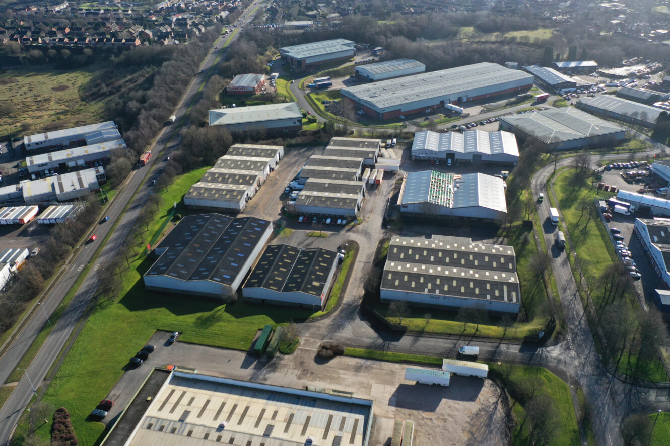 Aerial view of an industrial estate with multiple warehouses, access roads, parked vehicles, and surrounding greenery. Residential area visible in the background.