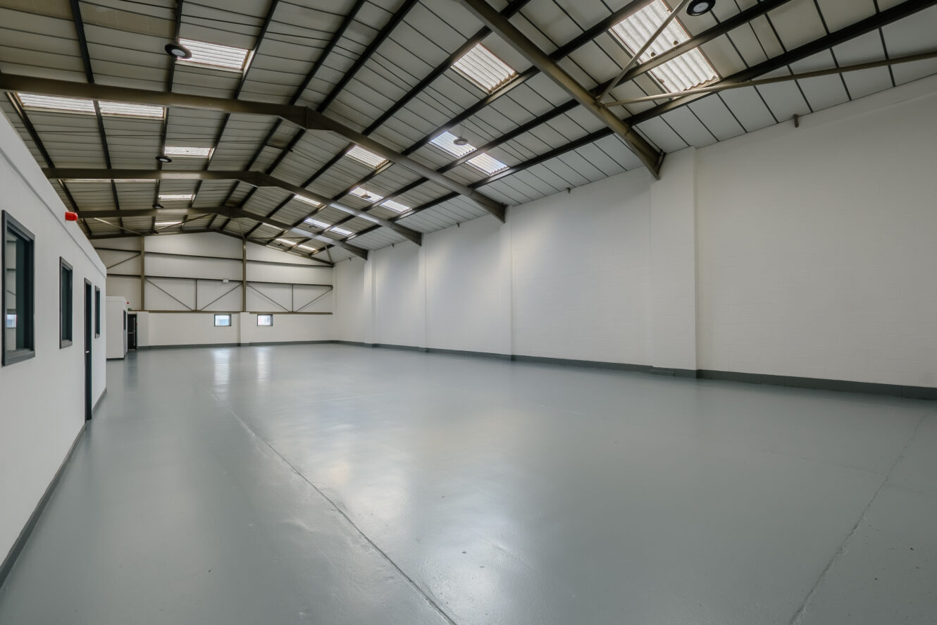 Empty industrial warehouse with high ceilings, white walls, gray floor, and several windows along the side and end walls.