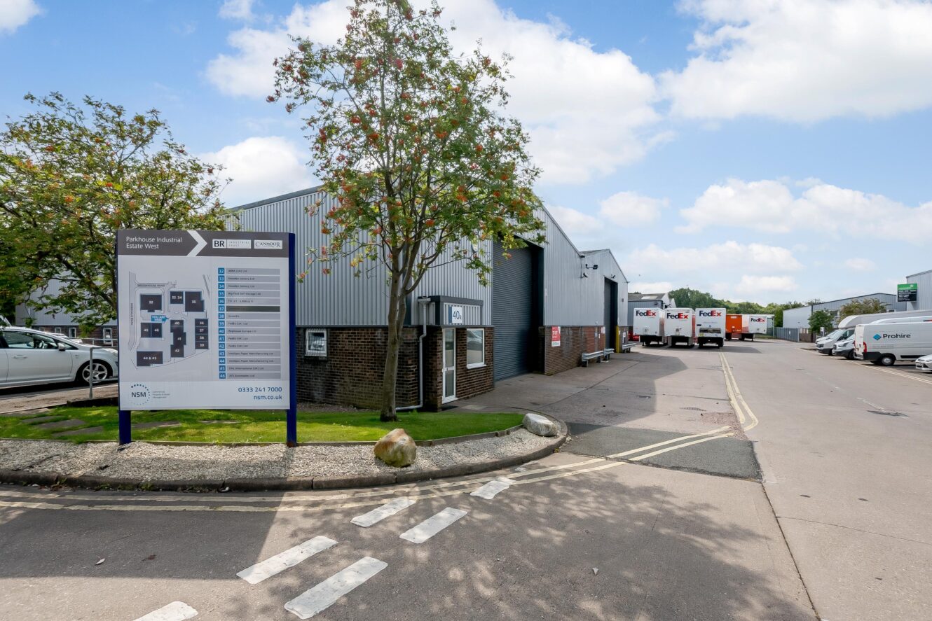 Industrial warehouse complex with a site map sign in the foreground, trees, and several FedEx trucks parked near loading bays in the background.