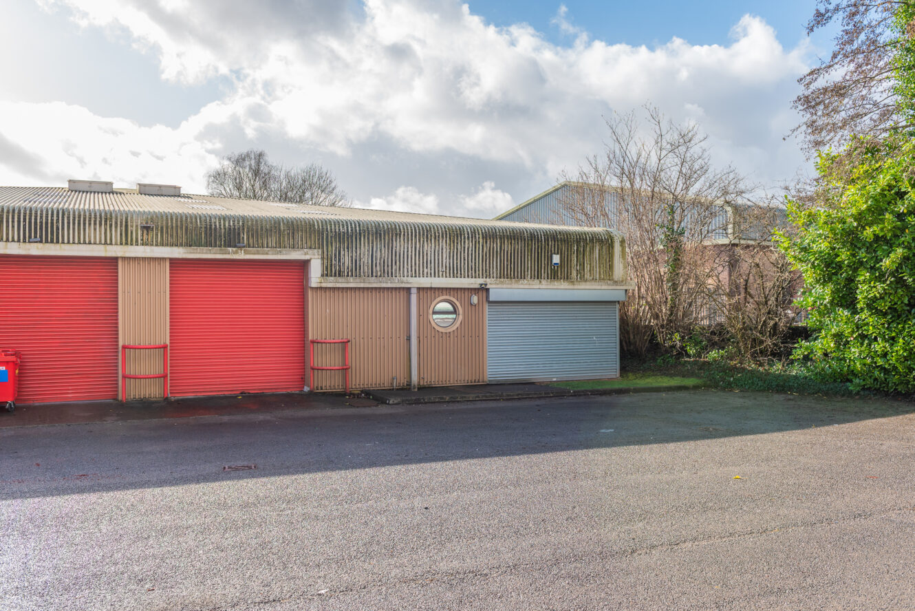 Single-story industrial building with two red roller doors, one gray roller door, a circular window, and an asphalt parking area in front. Trees and bushes are visible to the right.