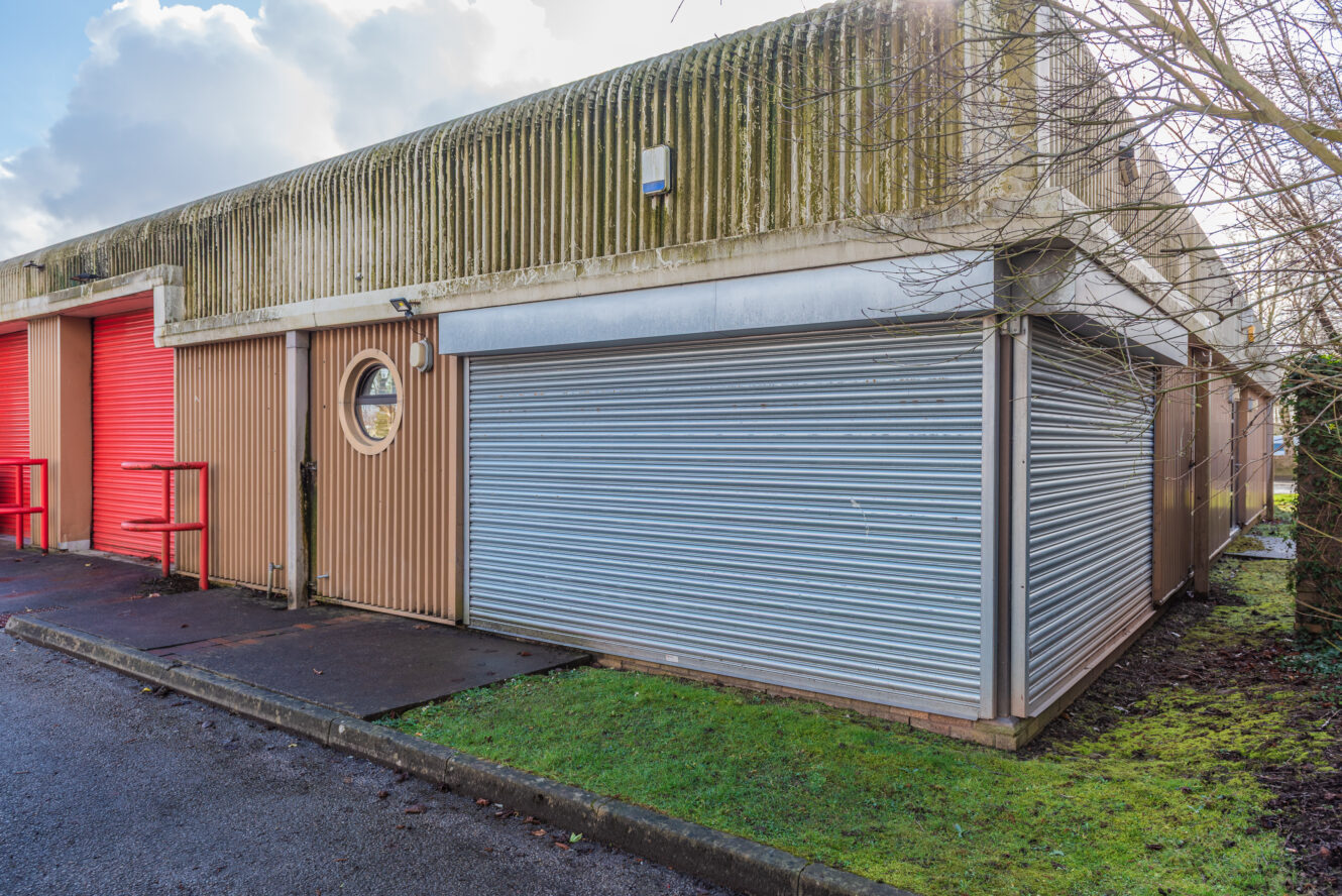 A concrete and metal industrial building with closed red and silver roller shutter doors, next to a patch of grass and wet pavement.