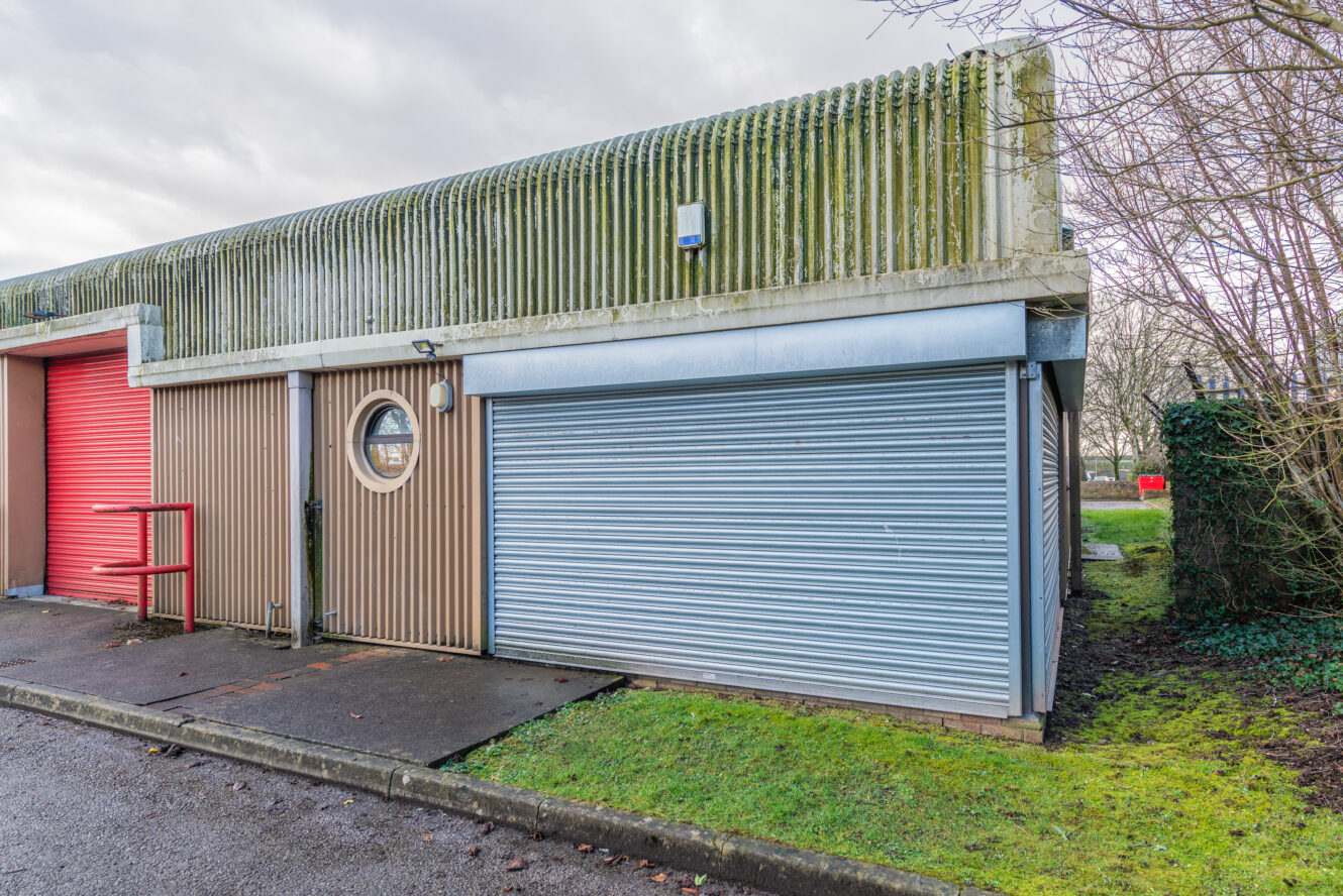 A small industrial building with a red door, blue roller shutter, round window, and moss on the roof, next to a patch of grass and trees.