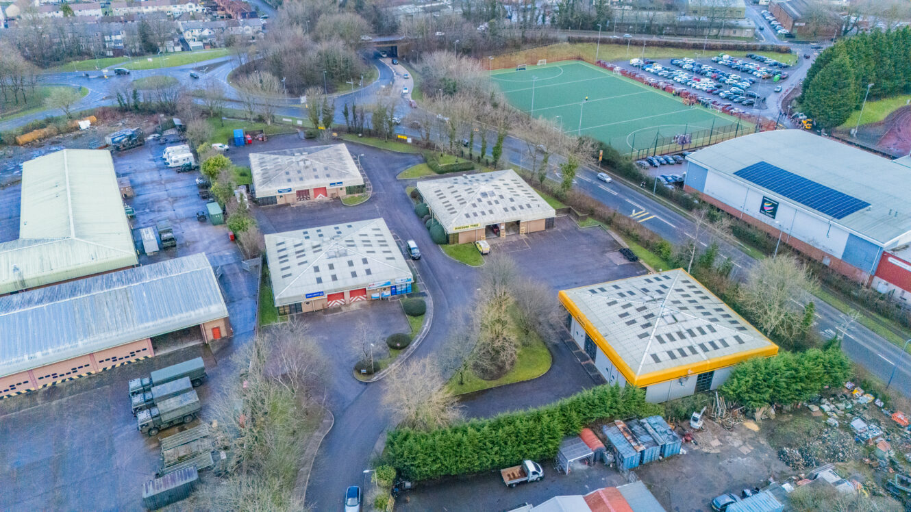 Aerial view of an industrial area with several warehouses, parking lots, a sports field, and surrounding roads lined with trees and parked cars.