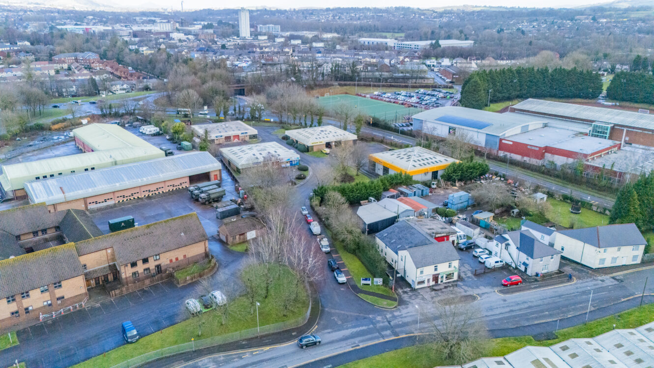 Aerial view of an industrial estate with warehouses, office buildings, parking lots, and roads surrounded by trees and residential areas.