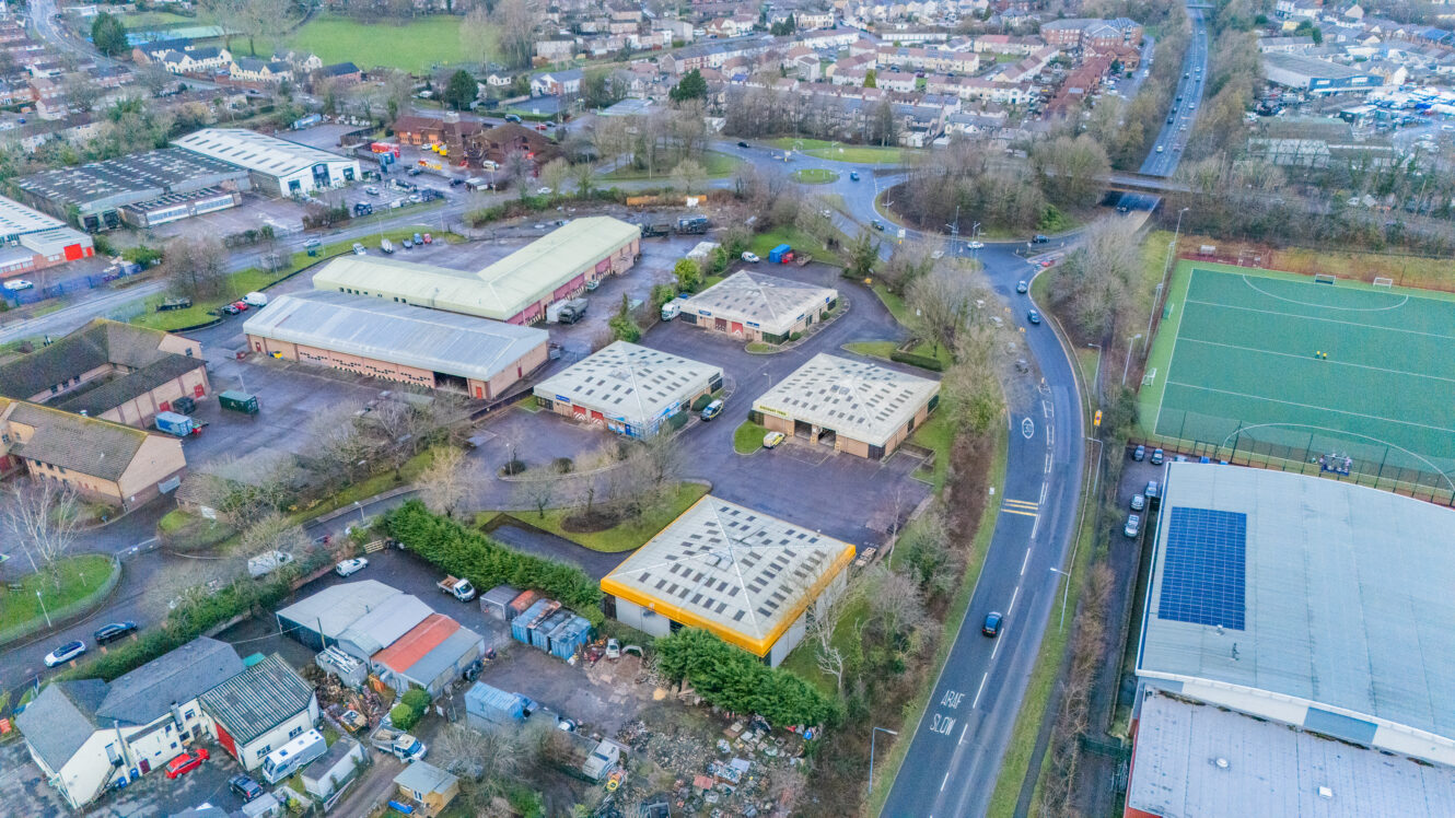 Aerial view of an industrial area with warehouses, surrounding roads, a sports field with solar panels, and residential houses in the background.