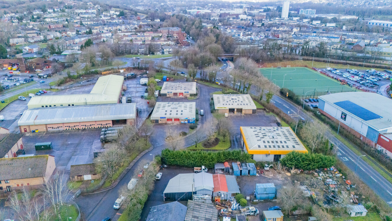 Aerial view of an industrial estate with multiple warehouses, a sports field, parking lot, and surrounding roads, with a town and hills in the background.