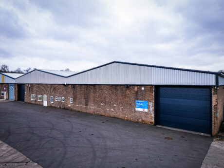 A large industrial warehouse with a brick exterior, multiple windows, a blue roller door, and a To Let sign is visible under a cloudy sky.