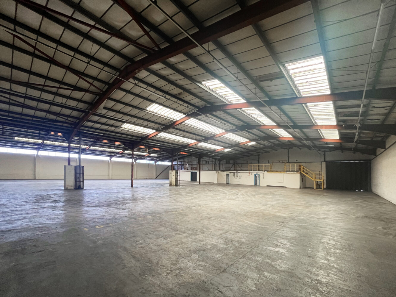 Spacious, empty warehouse with high ceilings, exposed beams, skylights, concrete floor, and a small mezzanine level with stairs on the right side.