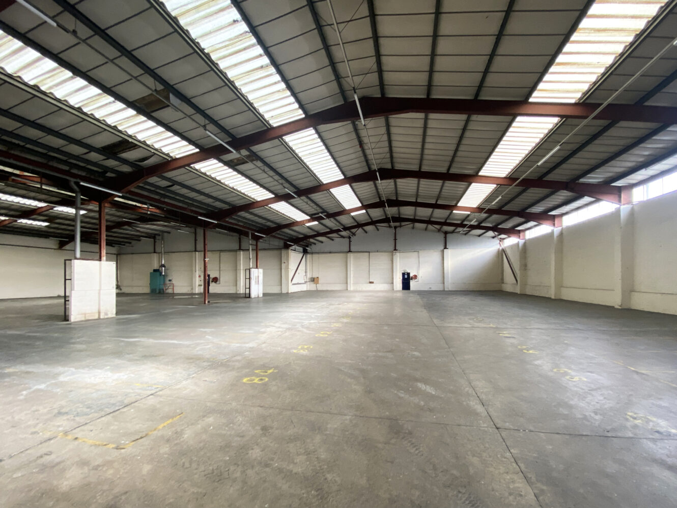 Interior of a large, empty warehouse with concrete floors, high ceilings, red steel beams, and skylights letting in natural light.
