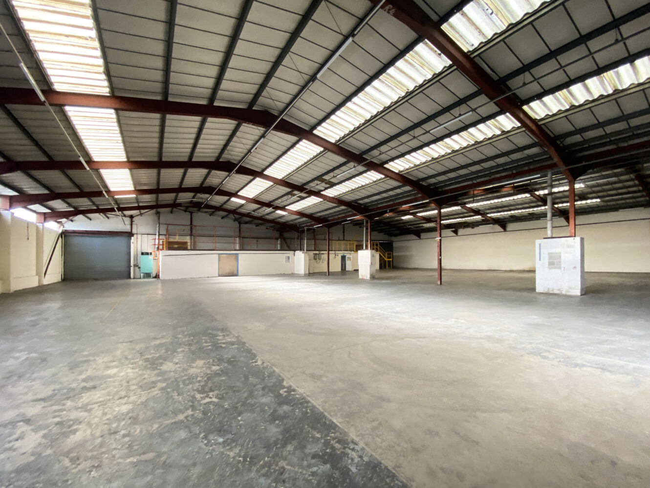 Large, empty warehouse interior with concrete floors, high metal roof, exposed beams, and walls lined with windows and doors.