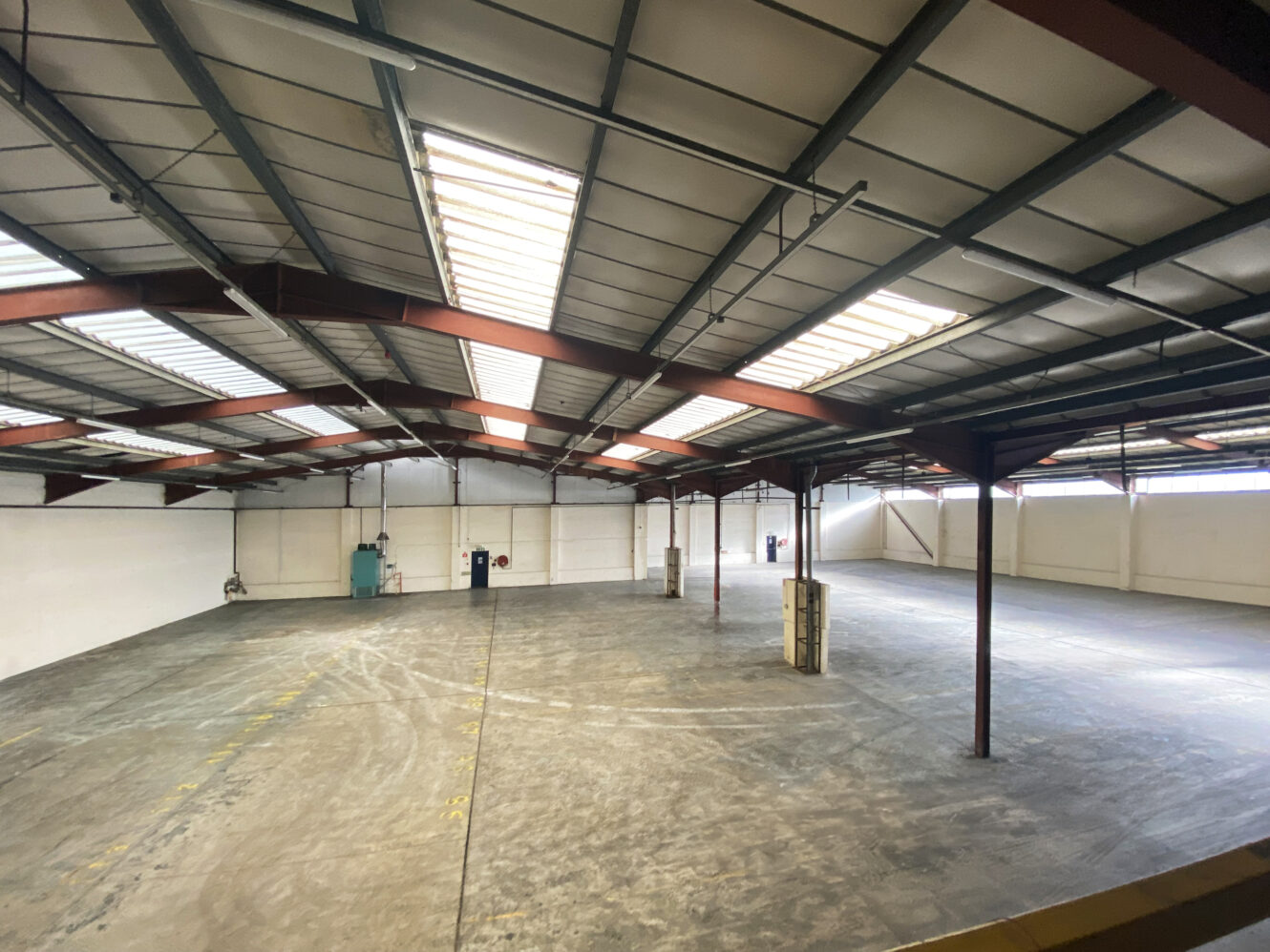 Interior view of a large, empty industrial warehouse with concrete floors, white walls, steel beams, and skylights in the ceiling.