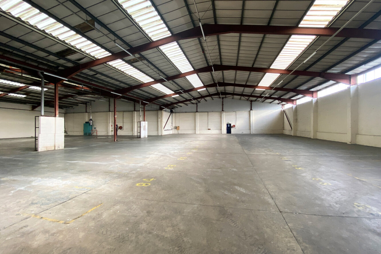 Spacious, empty warehouse with high ceilings, exposed beams, concrete floor, and large windows allowing natural light.