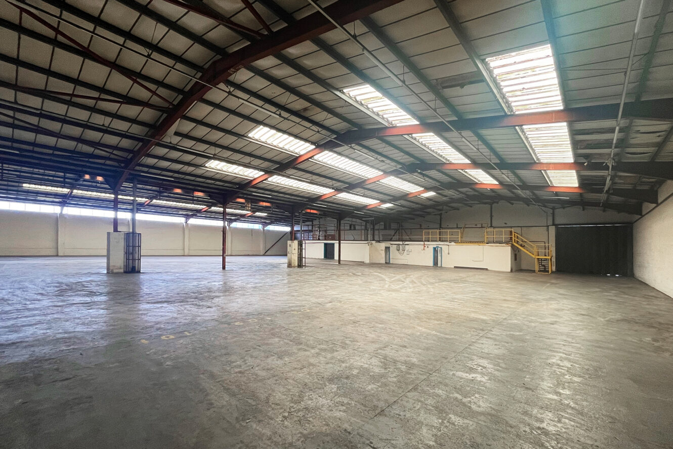 Large, empty warehouse with exposed steel beams, concrete floor, skylights, and a mezzanine level with yellow railings along the right side.
