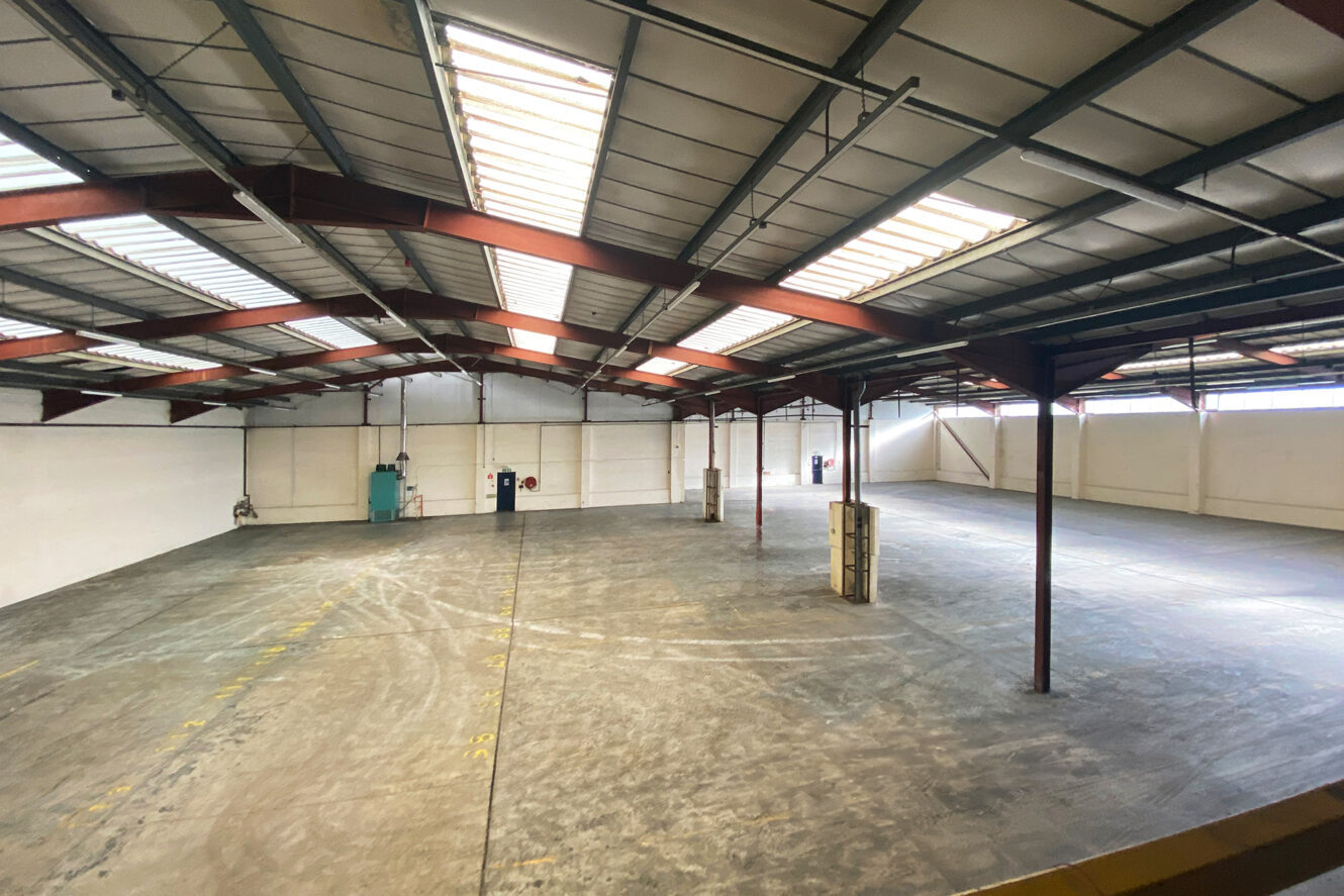 Interior view of an empty industrial warehouse with concrete flooring, steel beams, skylights, and minimal equipment along the white walls.