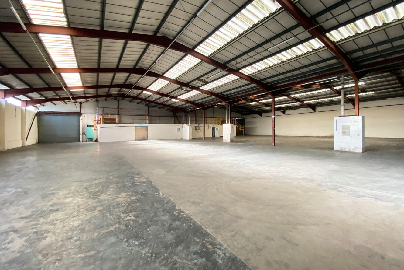 Interior of an empty warehouse with high ceilings, exposed beams, large windows, and a concrete floor. A mezzanine and closed office areas are visible in the background.