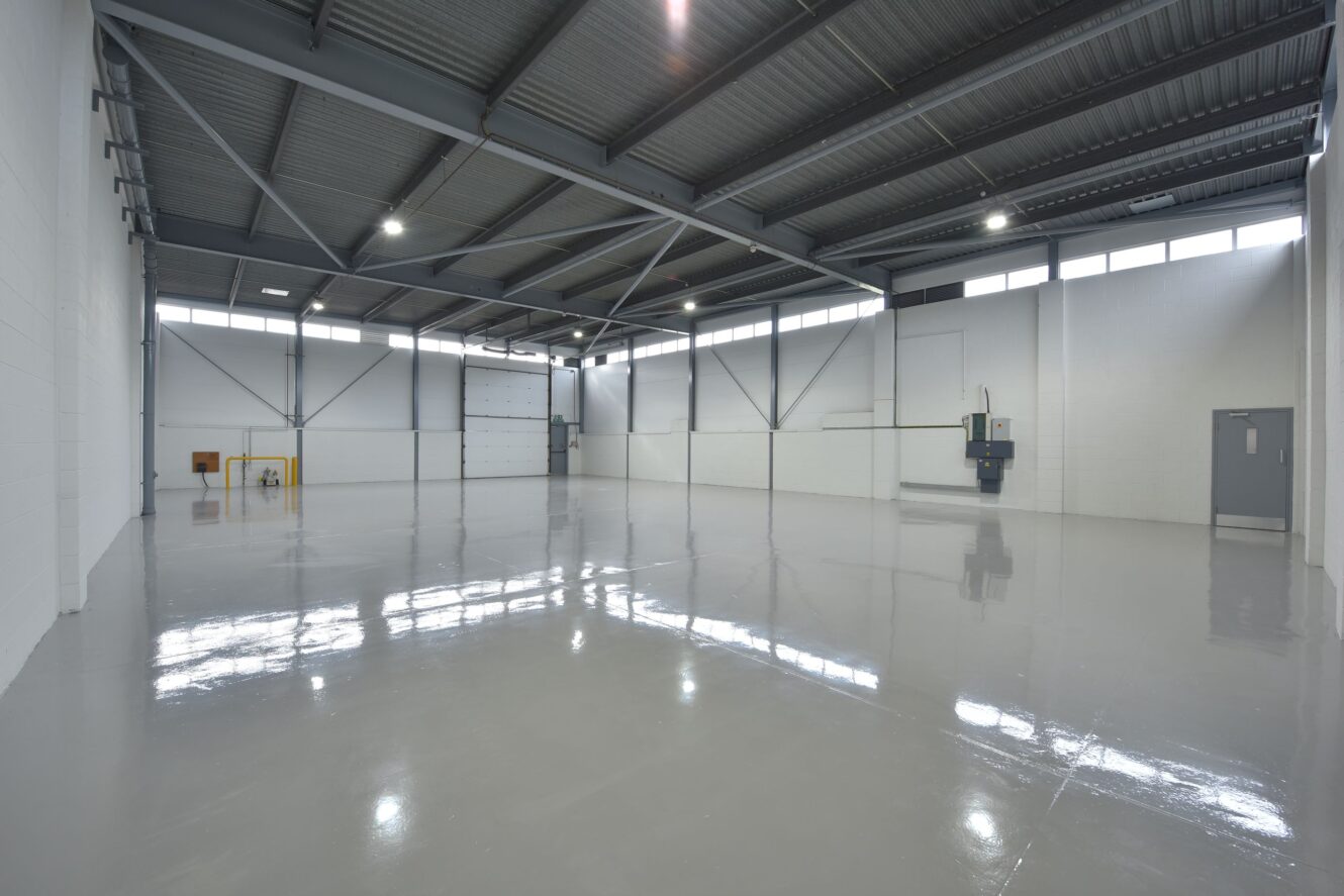 Large, empty industrial warehouse with polished concrete floors, high ceilings, exposed beams, and overhead lighting. Several doors and utility units line the white-painted walls.