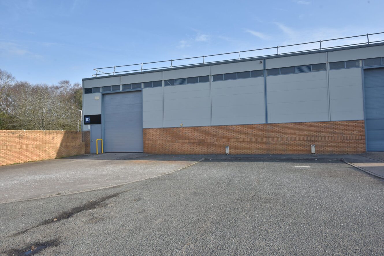 A large industrial building with brick and gray panel walls, two closed roller doors, and an empty paved parking area in front.