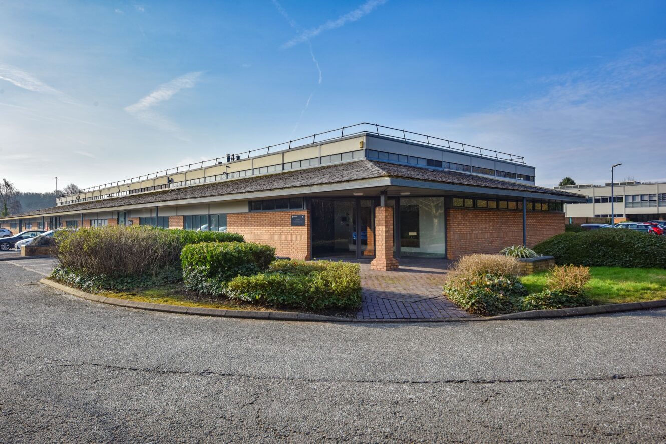 Single-story brick and glass office building with flat roof, surrounded by hedges and a parking lot under a clear blue sky.