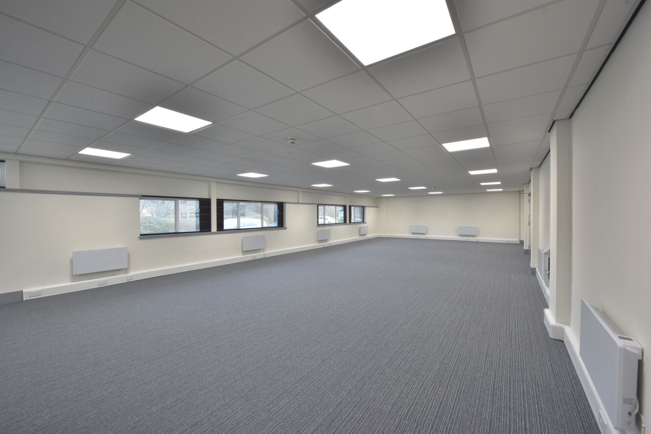 Empty office space with gray carpet, white walls, large windows, fluorescent ceiling lights, and wall-mounted heaters.