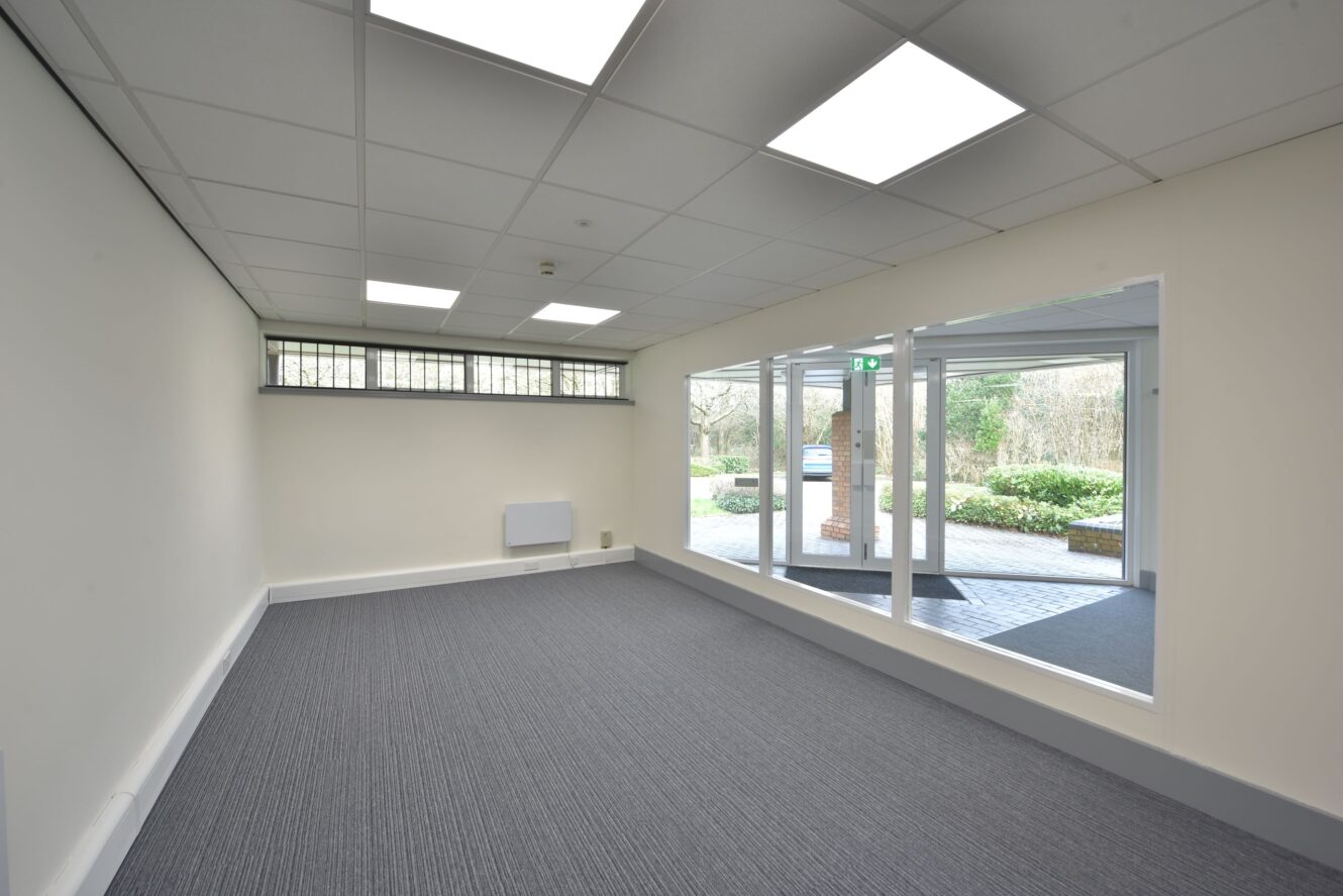 Empty office room with white walls, grey carpet, and large interior windows overlooking an outdoor area with greenery and brick columns. Fluorescent lights are on the ceiling.