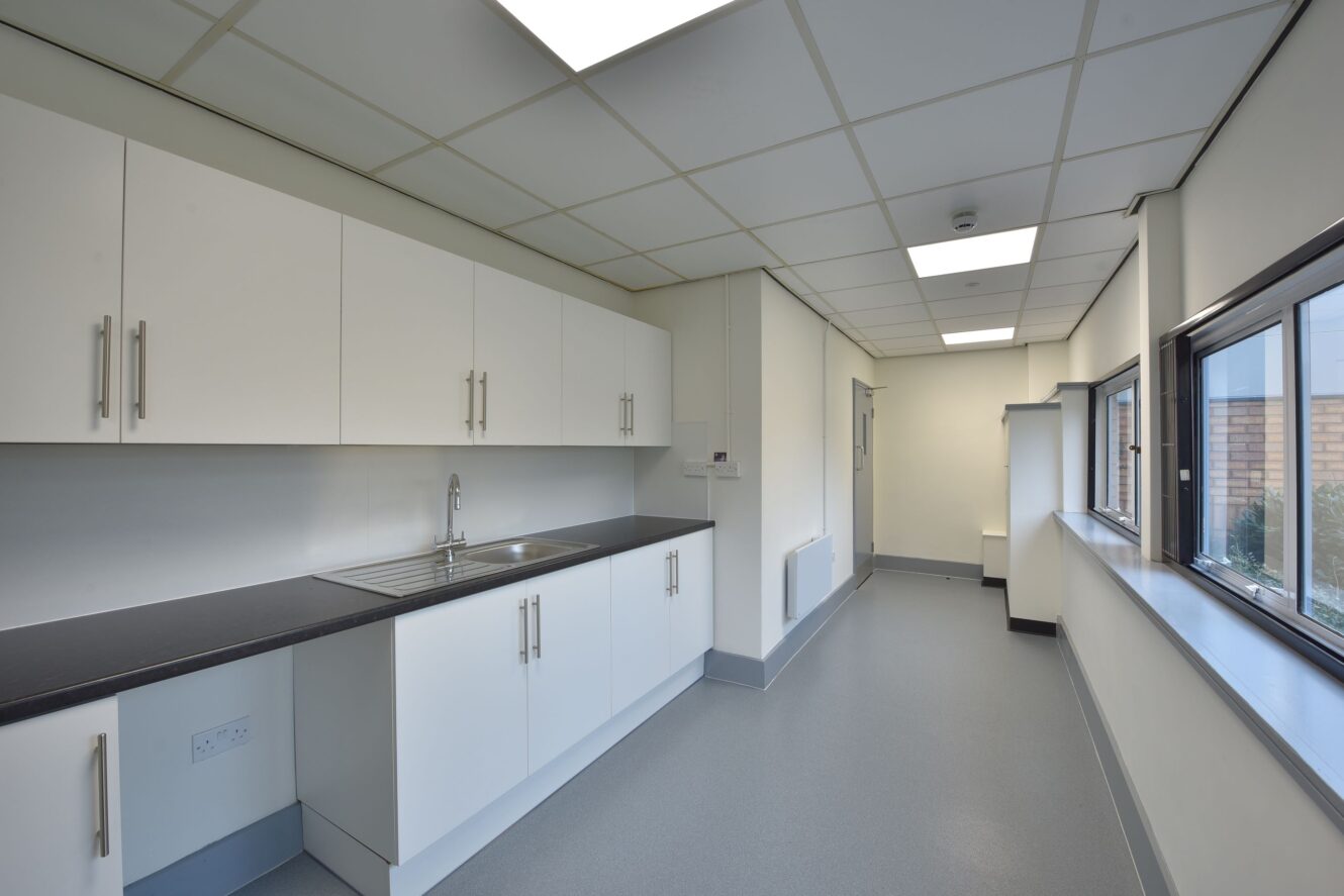 A clean, modern kitchenette with white cabinets, a stainless steel sink, black countertops, and large windows along one wall in a well-lit room.