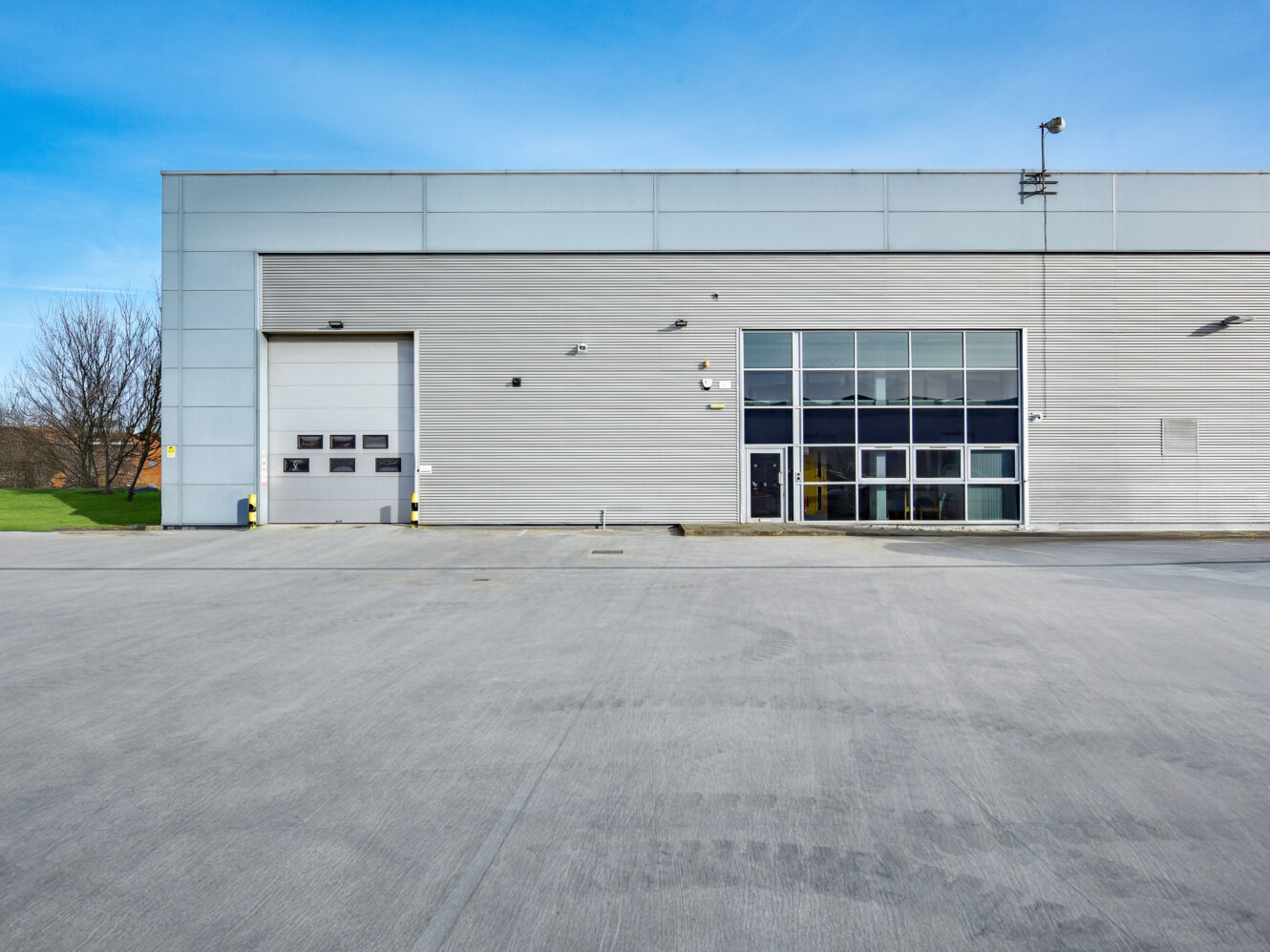 Exterior of a modern industrial warehouse with a large garage door, multiple windows, and a wide empty concrete lot in front.