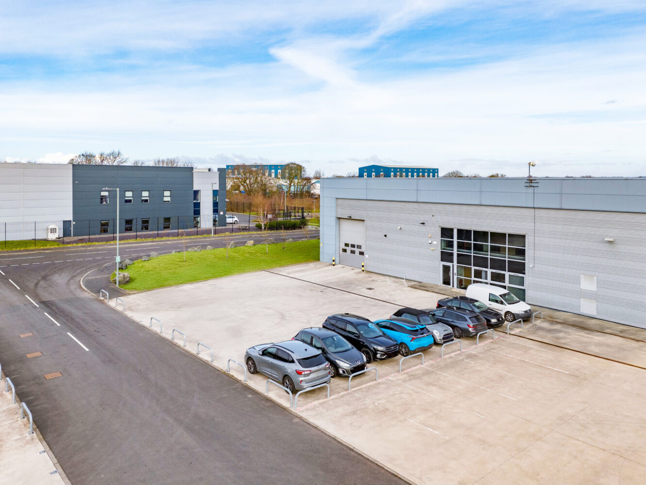 A row of cars is parked outside a large, modern industrial building with a wide driveway and adjacent road, under a partly cloudy sky.