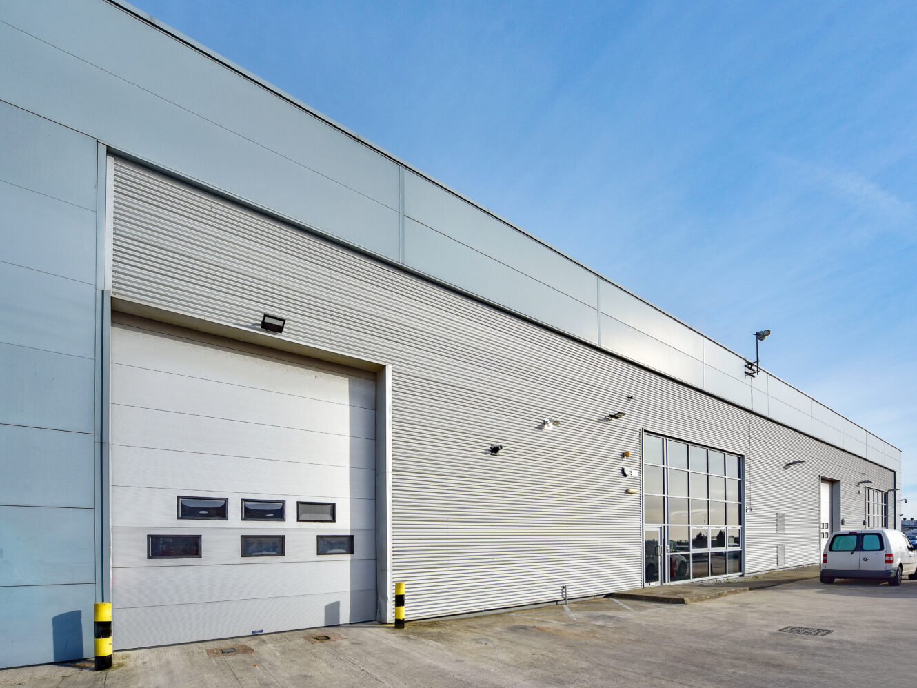 Exterior view of a large industrial warehouse building with a roller shutter door, glass windows, and a small white van parked nearby.
