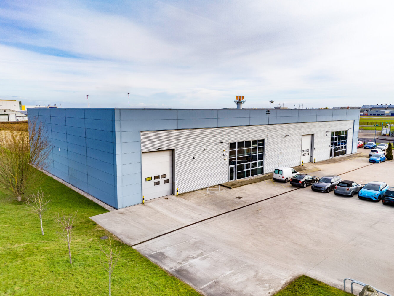 Large modern industrial warehouse with light blue and white exterior, several loading bays, and parked cars in the front lot on a clear day.