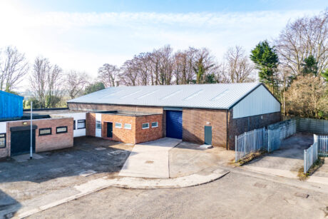 A brick industrial warehouse with a metal roof and blue roller door, surrounded by trees and fencing, photographed on a clear day.