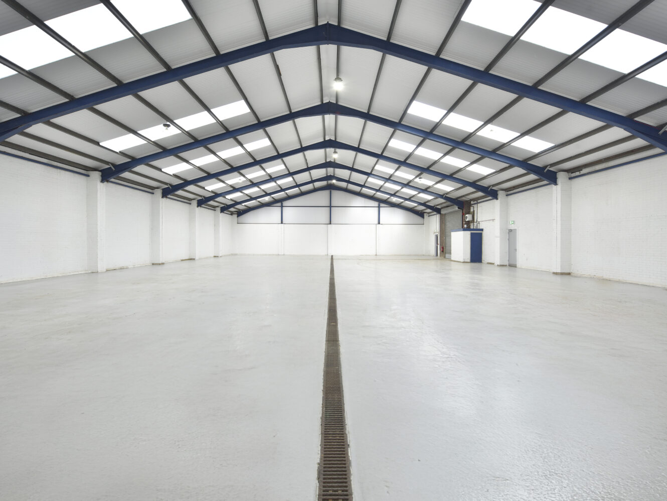 Empty industrial warehouse interior with white walls, high ceiling, and metal beams. The floor has a central drainage channel and the space is well-lit by skylights and windows.