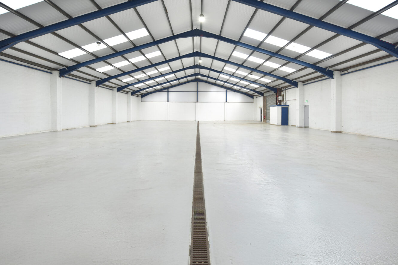Spacious, empty warehouse interior with white walls, a high ceiling, exposed beams, and a grated floor drain running down the center.