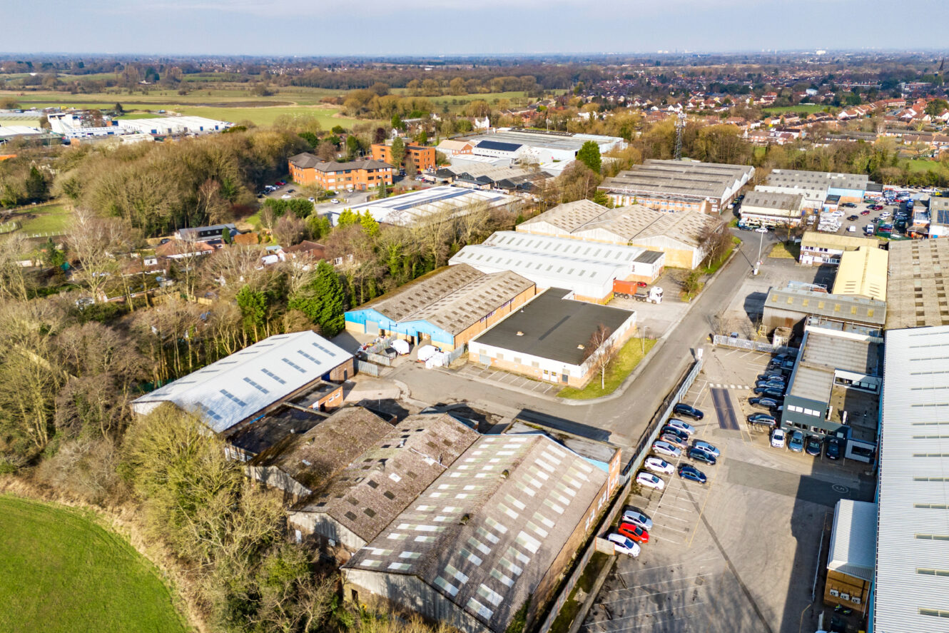 Aerial view of an industrial estate with warehouses, parking lots, and surrounding trees, with residential areas visible in the background.