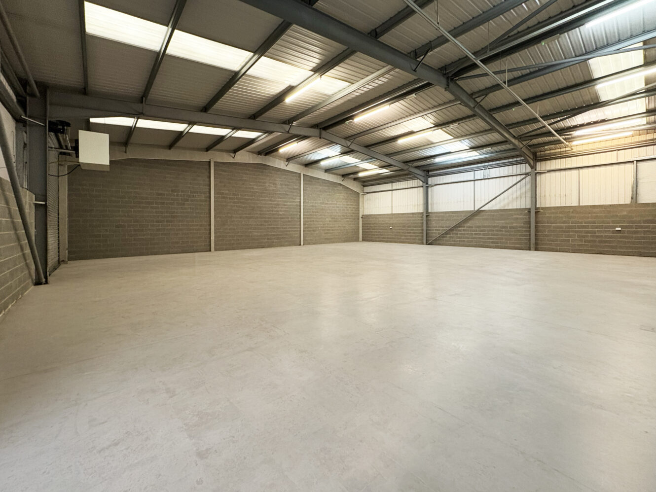 Large empty warehouse with concrete floor, brick walls, and metal roof featuring skylights and fluorescent lighting.