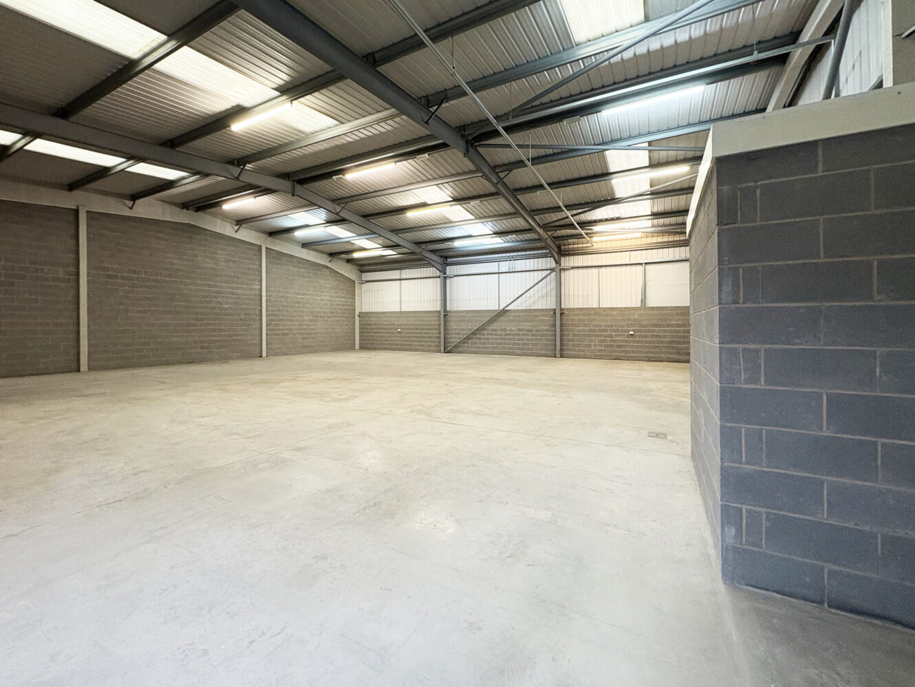 Empty industrial warehouse with concrete floor, brick walls, and metal ceiling, illuminated by overhead lights and skylights.
