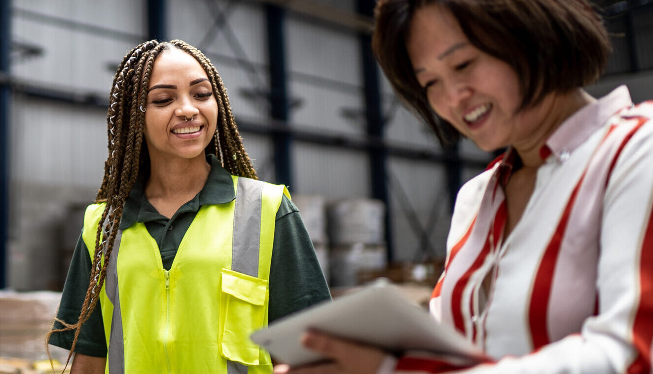 Two women in a warehouse, one wearing a yellow safety vest and the other holding a tablet, are having a discussion.