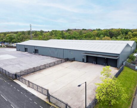 Large industrial warehouse with a grey exterior, loading bays, and spacious fenced concrete yard, set beside a road and surrounded by trees.