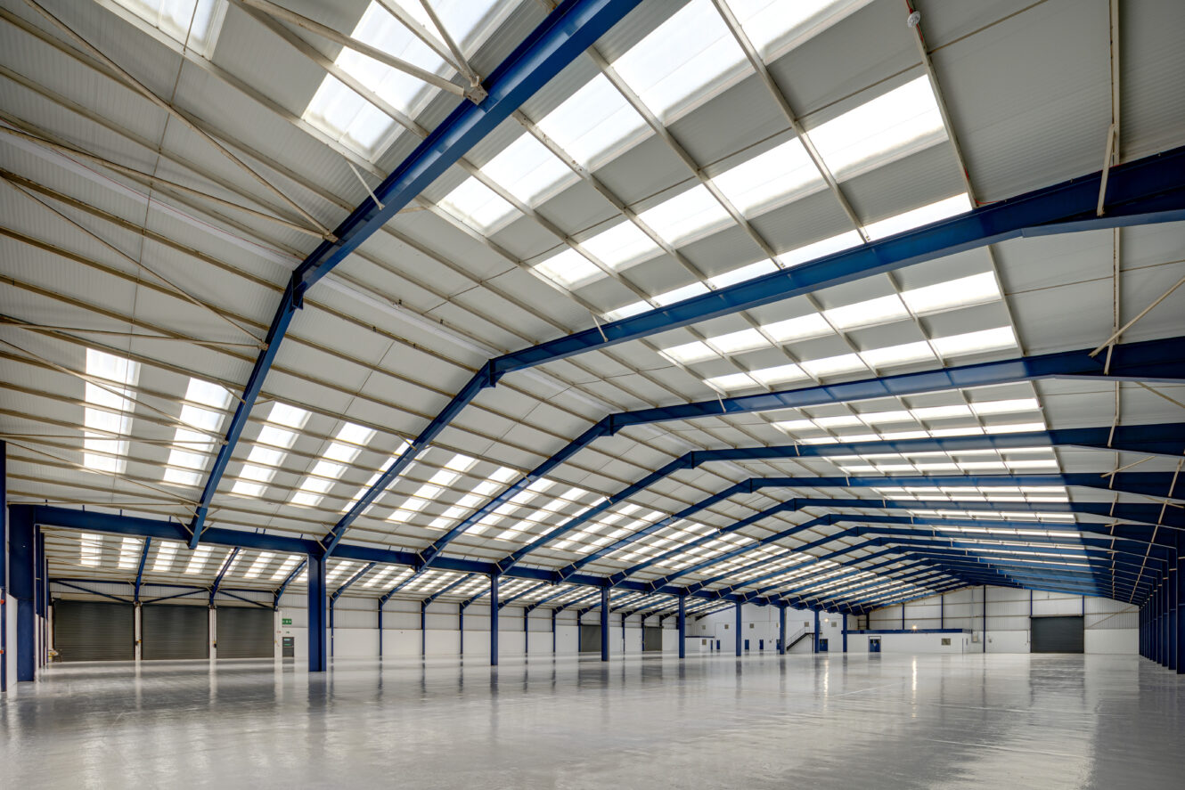 Interior of a large, empty warehouse with high ceilings, blue steel beams, and skylights allowing natural light to fill the space.