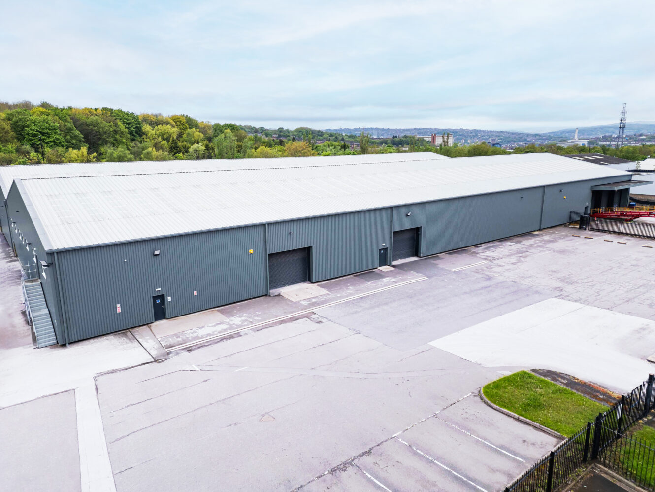 Large grey industrial warehouse with multiple loading bays, surrounded by an empty paved lot and greenery in the background under a cloudy sky.