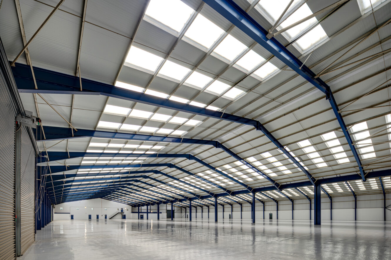 Large empty warehouse with high ceilings, skylights, and blue steel beams; polished concrete floor and white walls with multiple doors visible.
