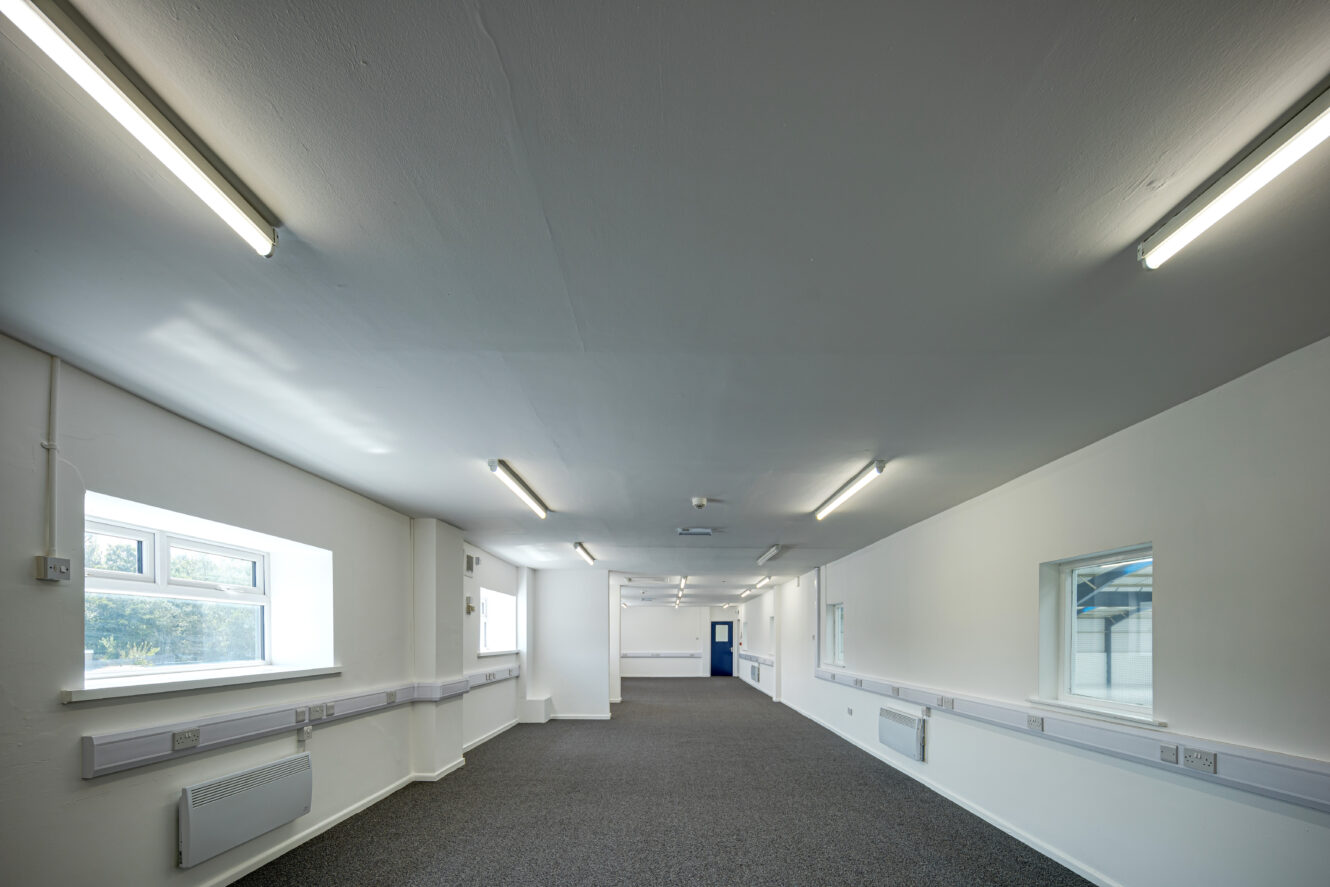 A long, empty office space with white walls, fluorescent ceiling lights, grey carpet, and several windows on both sides.