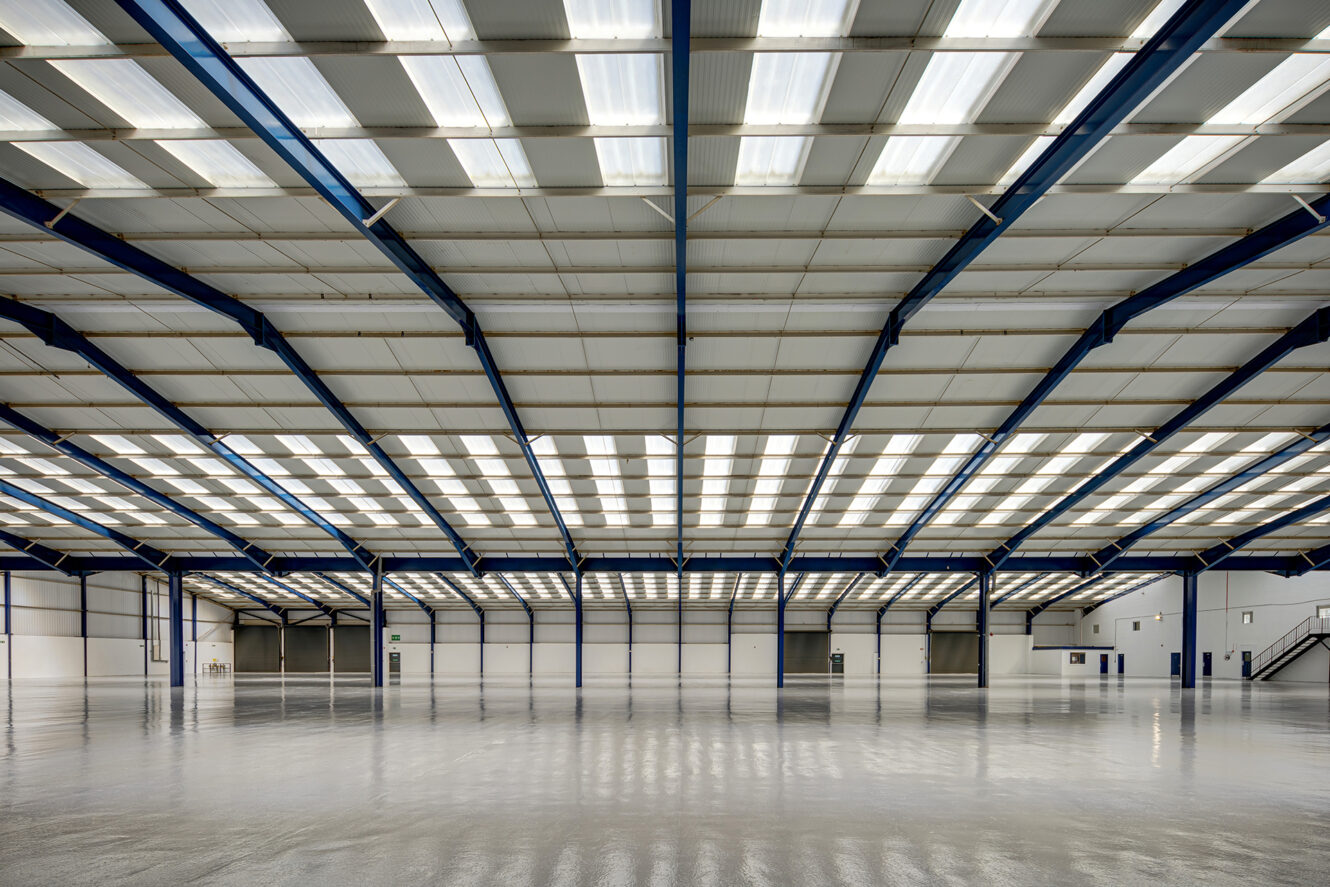 Interior view of a large, empty warehouse with a polished floor, high ceiling, exposed beams, and fluorescent lighting.