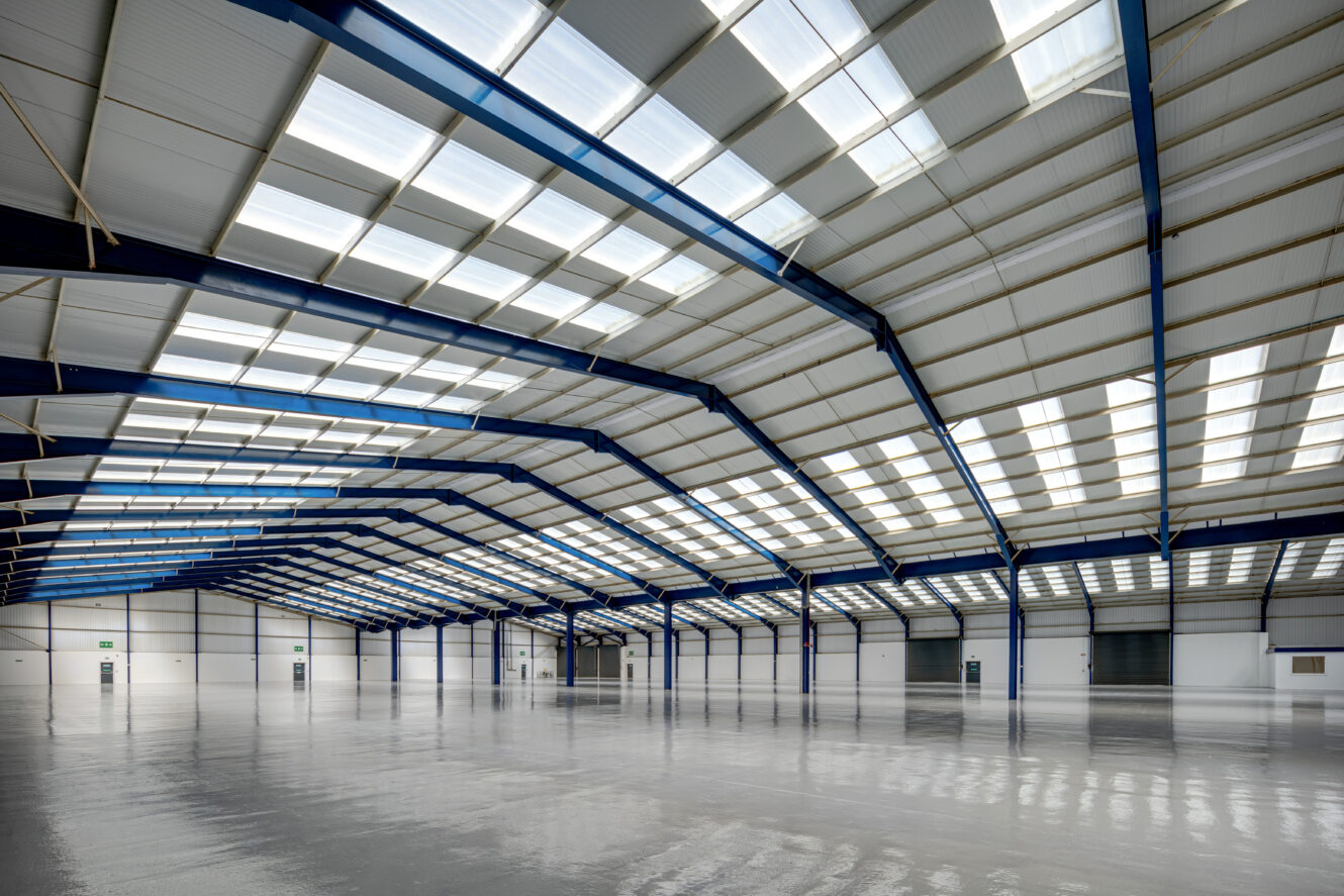 Large, empty warehouse interior with high ceilings, blue steel beams, and rows of skylights allowing natural light onto the polished concrete floor.