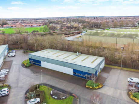 Aerial view of a rectangular industrial warehouse with a blue and gray exterior, surrounded by parking spaces and landscaped areas, with nearby buildings and a sports field in the background.