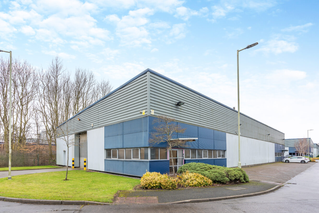 A modern industrial warehouse with metal siding, large windows, and a small landscaped area in front, situated on a paved lot under a partly cloudy sky.