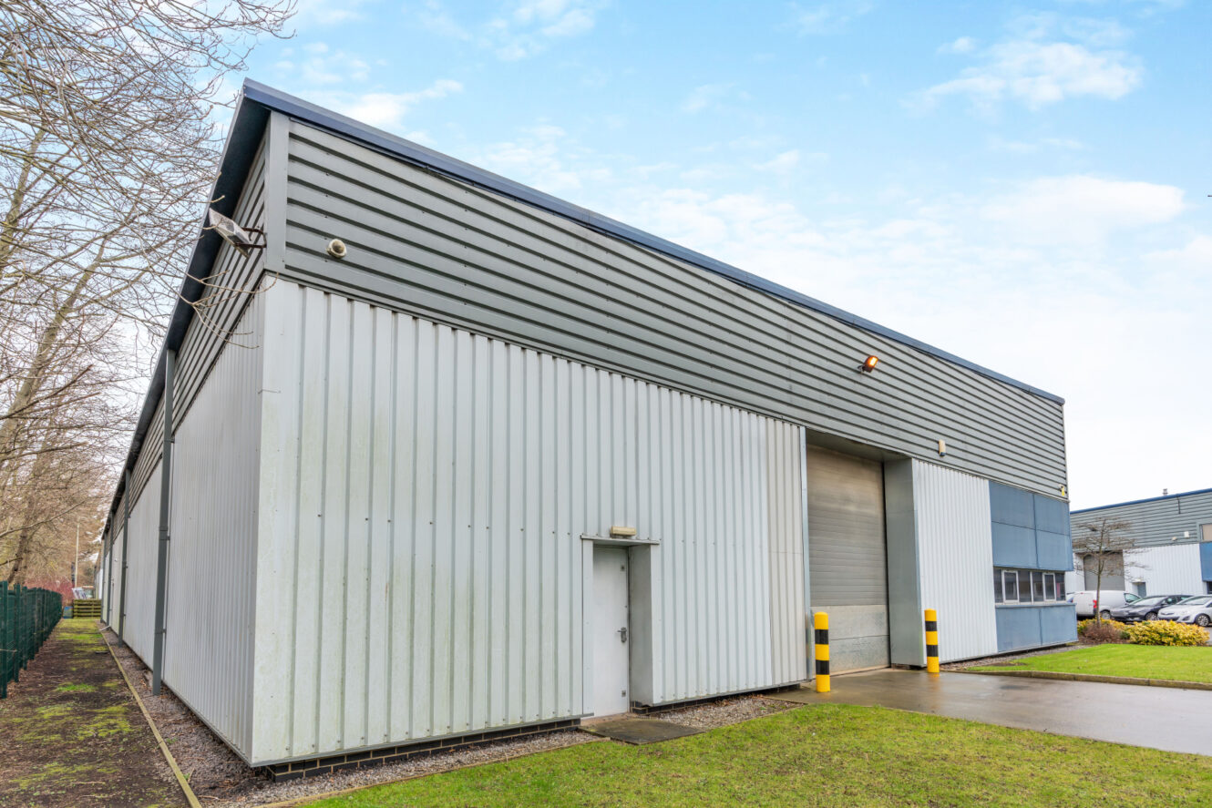 A large, modern industrial warehouse with metal siding, a small entrance door, and a large roller door, set on a grassy plot with a clear sky above.