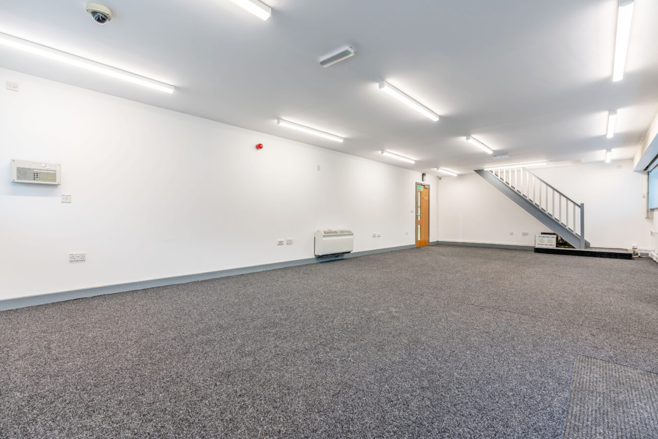 Empty office space with gray carpet, white walls, fluorescent ceiling lights, a staircase, and a wooden door.