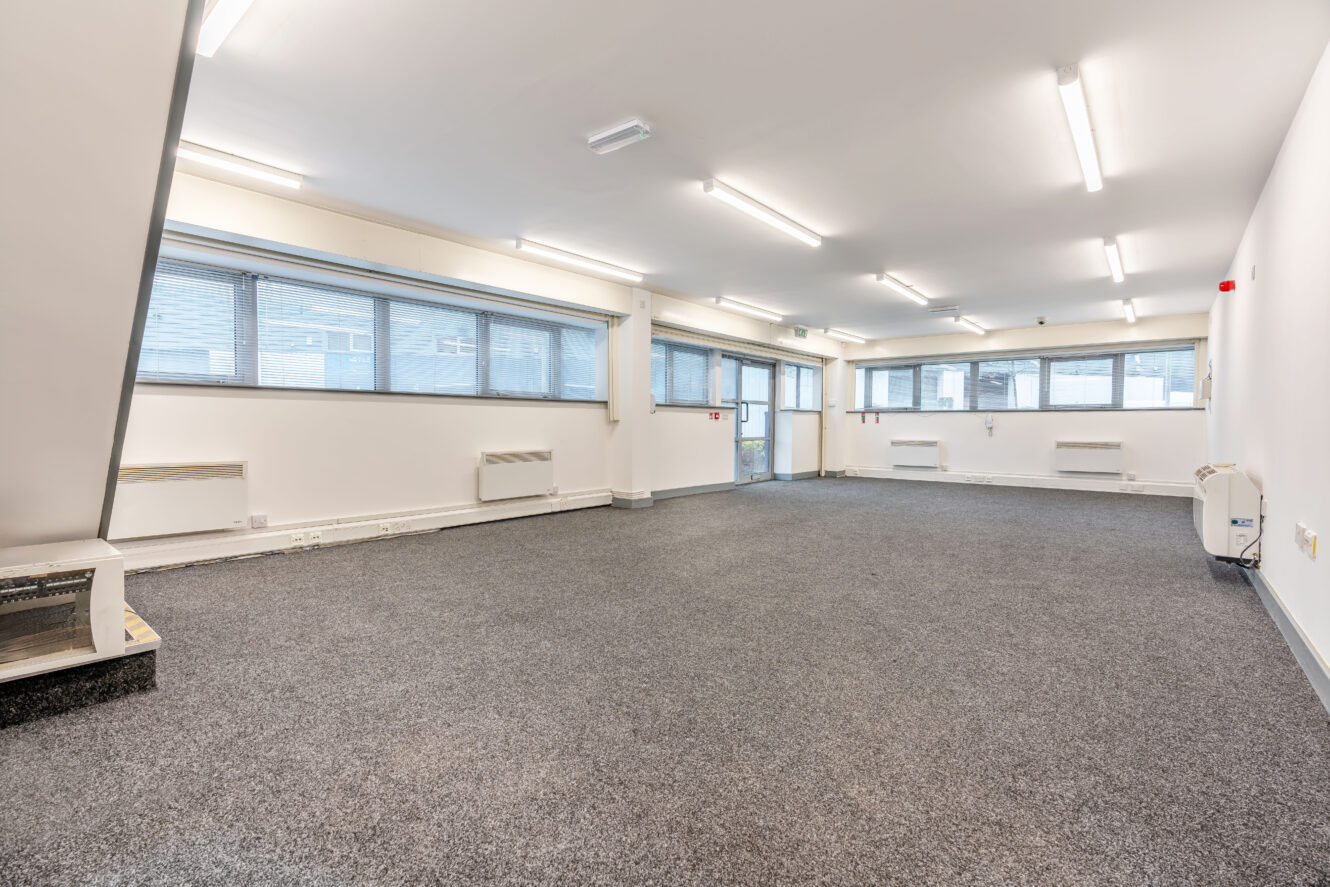A spacious, empty office room with grey carpet, white walls, large windows along one side, and fluorescent ceiling lights.