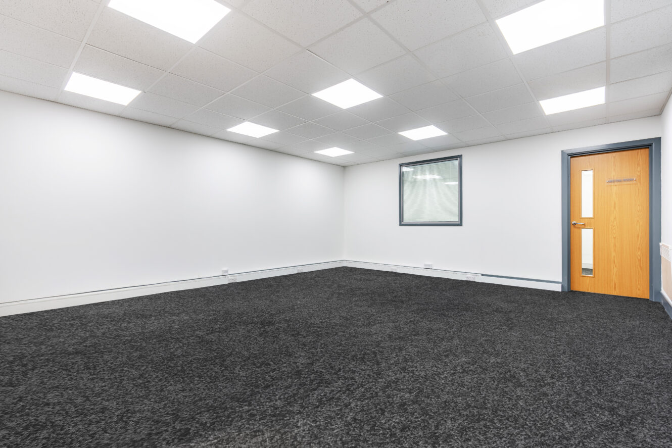 Empty office room with white walls, grey carpet, ceiling lights, a single window, and a wooden door with a glass panel.